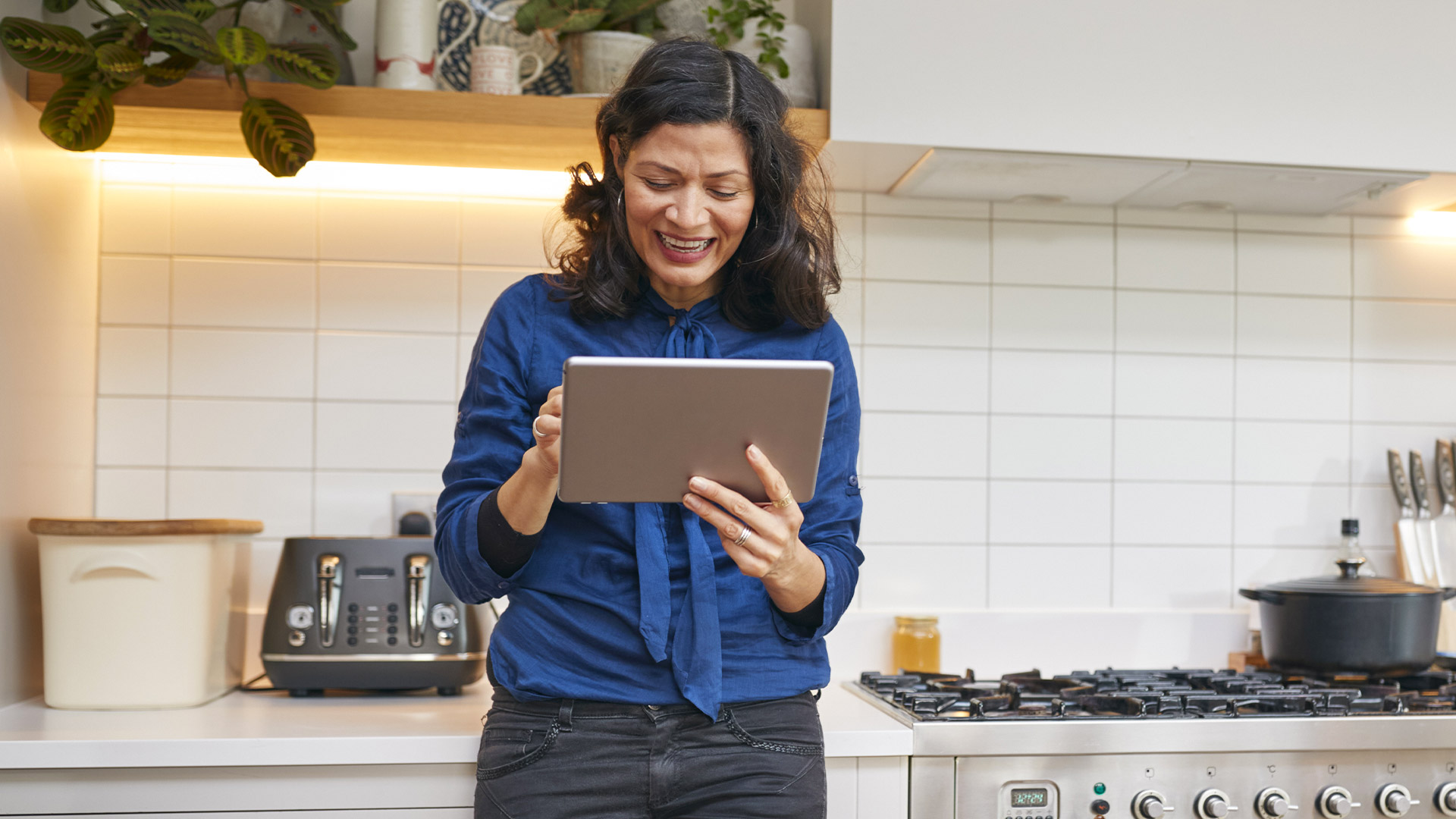 A woman with dark hair wearing a blue blouse stands leaned against her kitchen counter holding a tablet, smiling at the screen. 