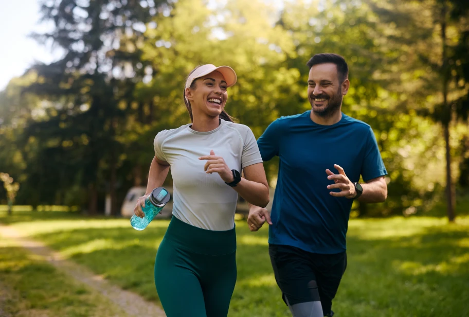 Two people jog on outdoor path, one wearing white t-shirt with cap holding blue water bottle, one wearing blue shirt, with trees in background