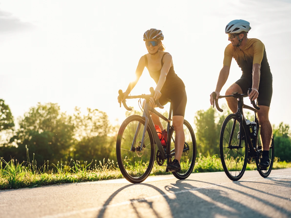 Two cyclists on road bikes wearing helmets and cycling shorts ride on paved path in bright sunlight with trees in background