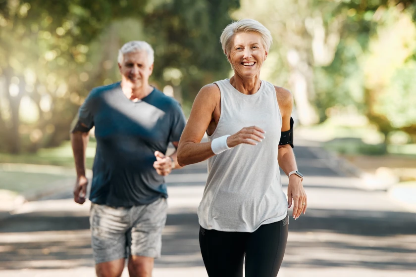 Two people jog side by side outdoors on paved path, one wearing navy blue athletic top and one wearing light gray athletic top