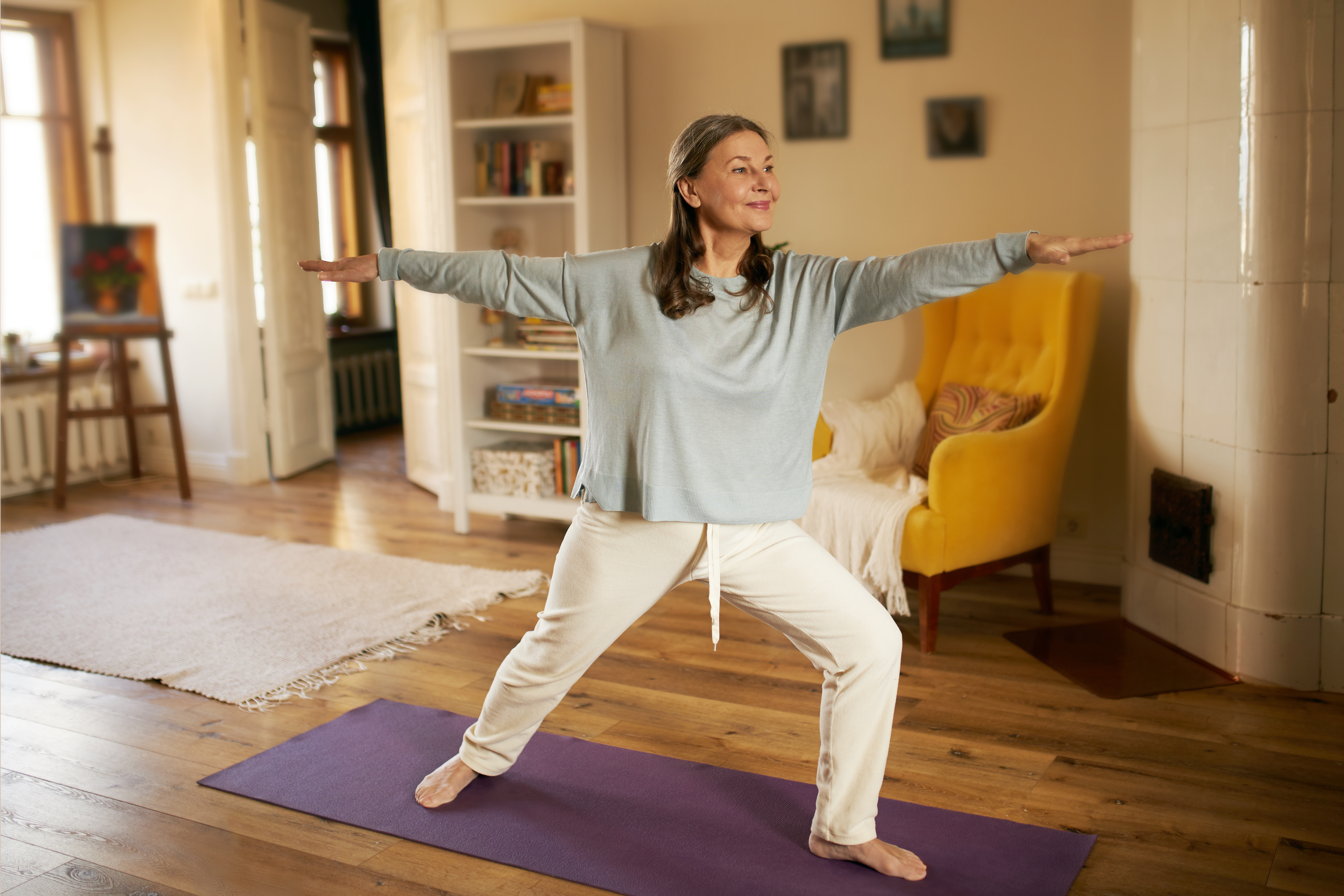 Middle aged woman in living room standing and stretching on Yoga Mat 