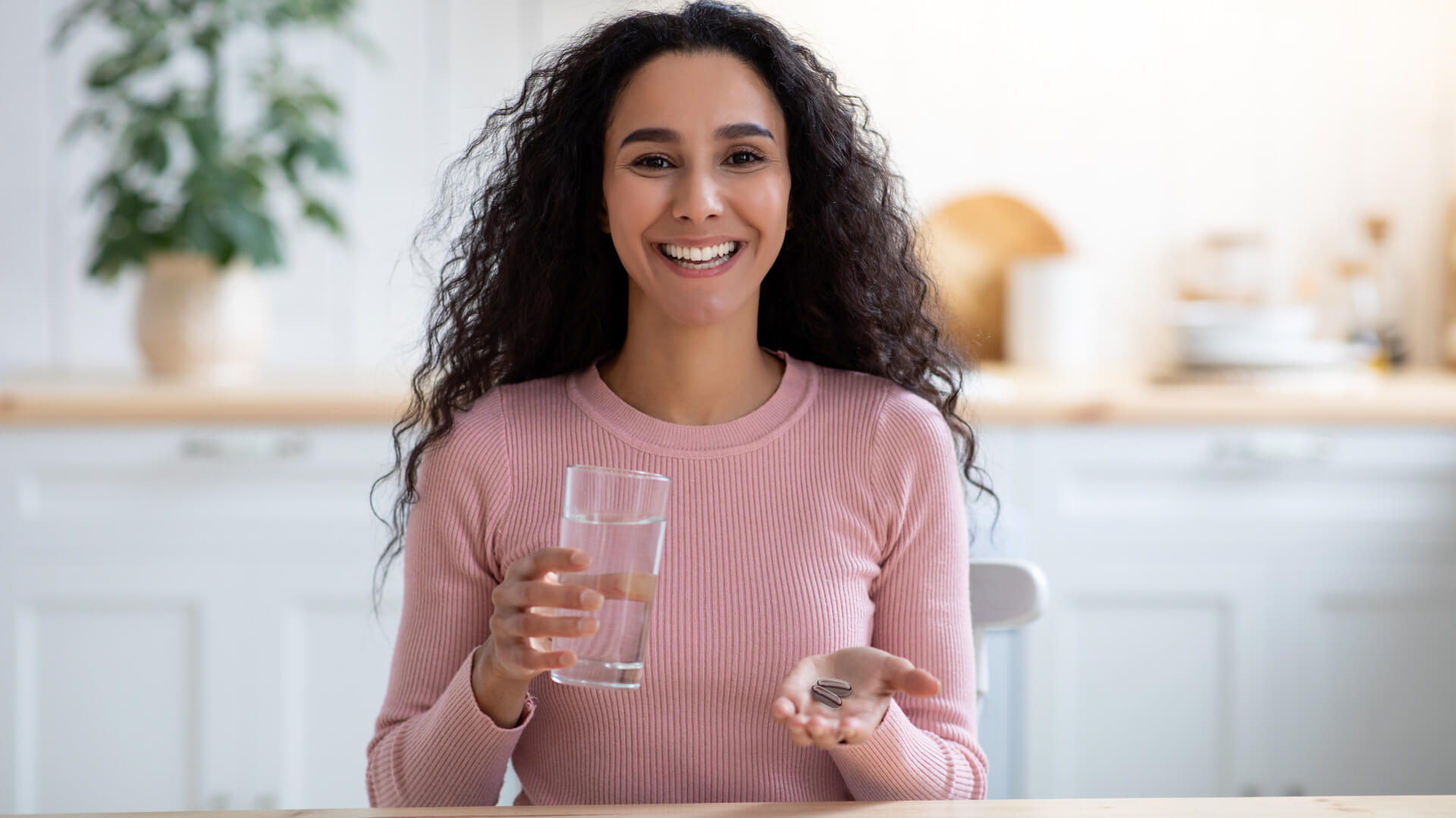 Woman in kitchen smiling with glass of water and 2 pills in hand