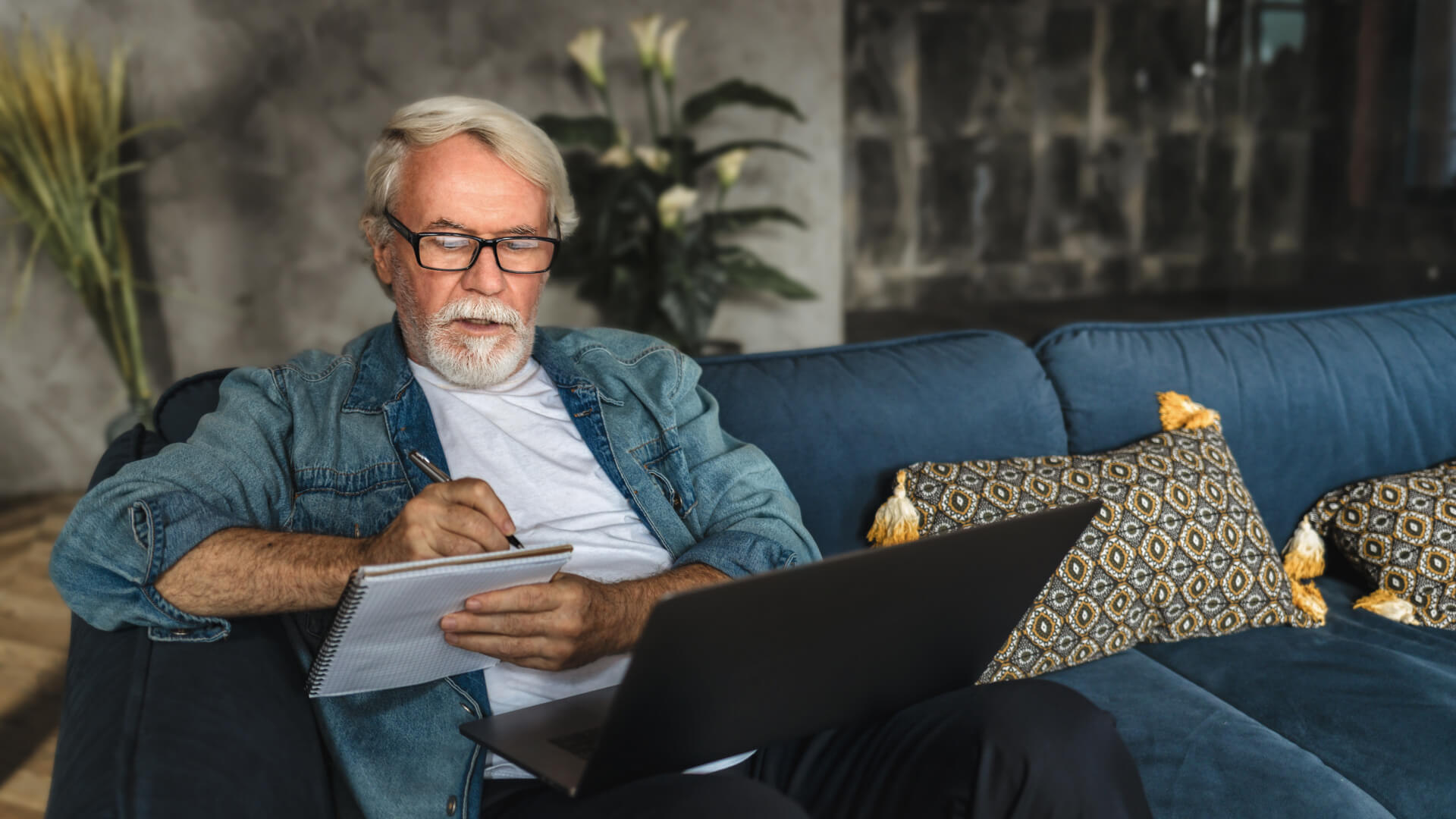 Senior man with glasses sitting on couch with notepad and laptop in lap