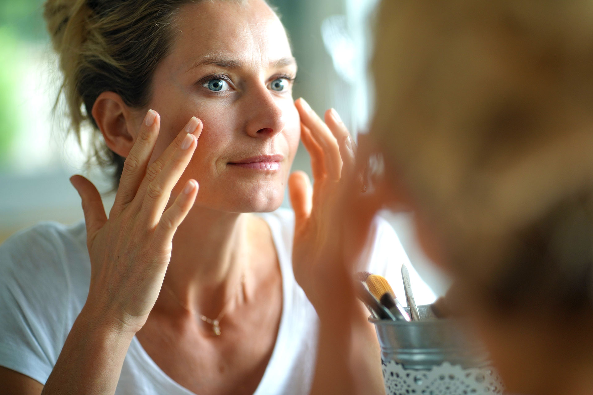 A woman looks in the mirror and carefully applies a skincare product to her cheekbones.