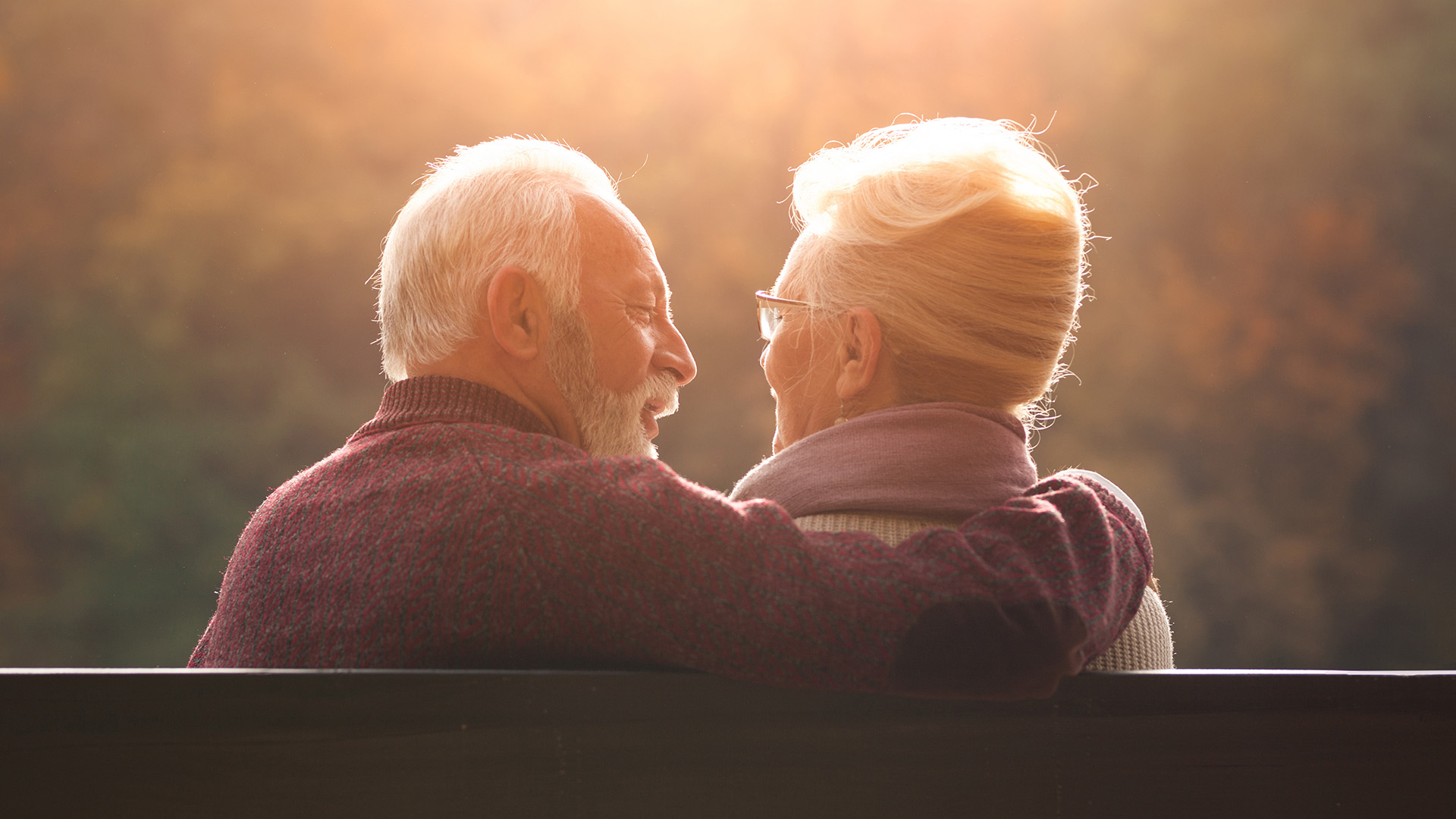 Senior couple sitting outside on bench with man's arm around woman in evening sun