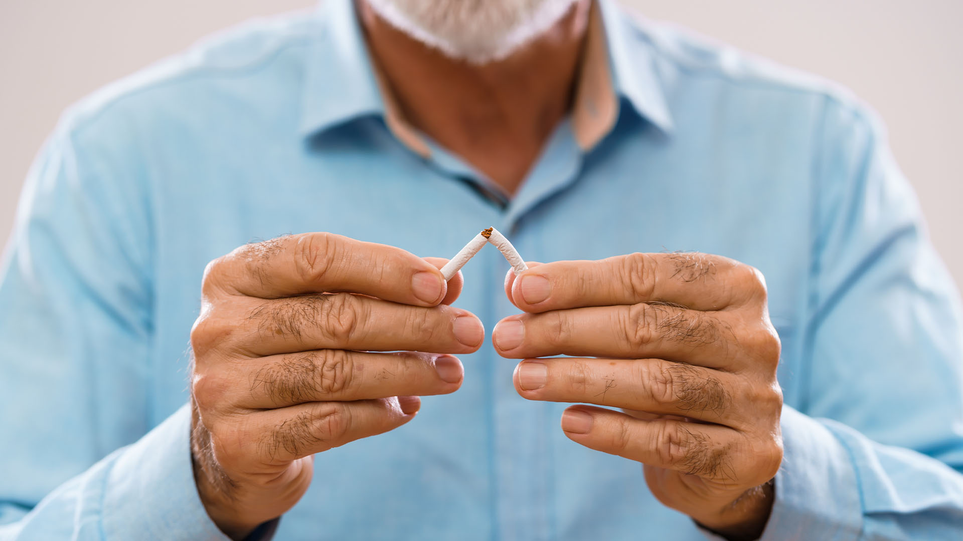 Man with white beard in Blue button up shirt, breaking a cigarette in half with his hands