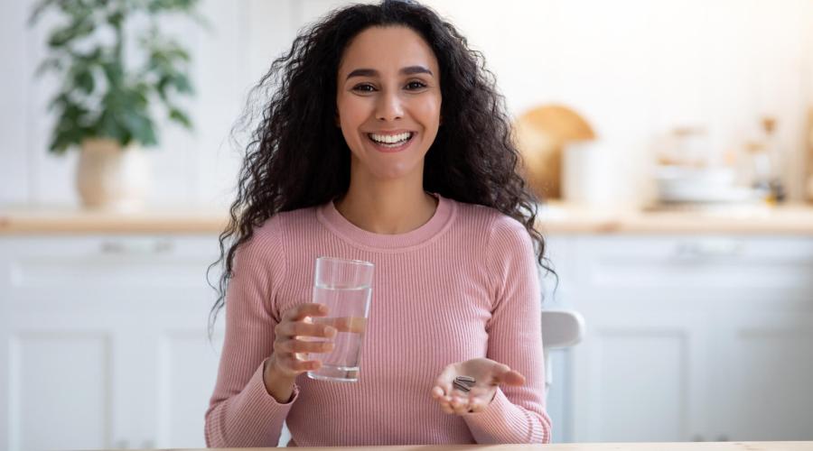 Woman in kitchen smiling with glass of water and 2 pills in hand