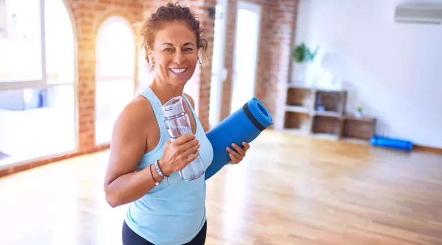 Middle-aged women smiling in athletic wear, holding water bottle and Yoga mat