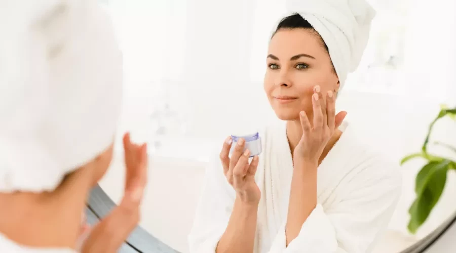 Woman in bathroom mirror with towel on her head and holding a small jar with one hand and applying to cheek with other hand