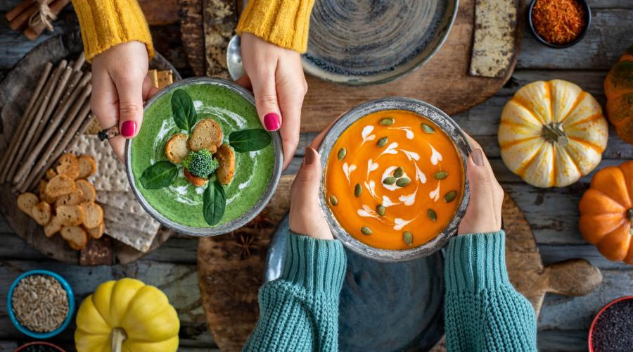 Womens hands holding bowls of vegetable soup over a wood table displayed with pumpkins, vcrackers, spices and nuts