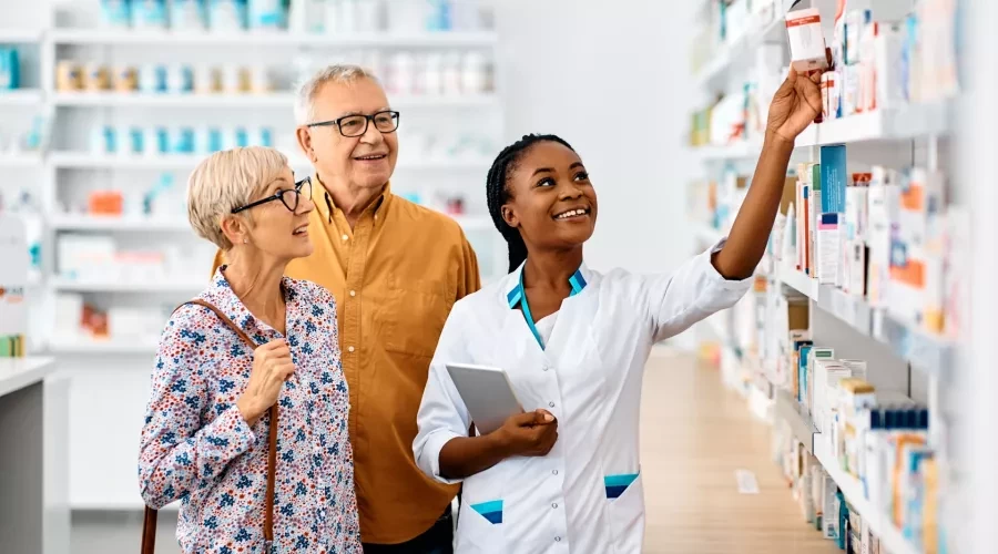 Pharmacist smiling and showing senior couple supplements on shelf in Pharmacy