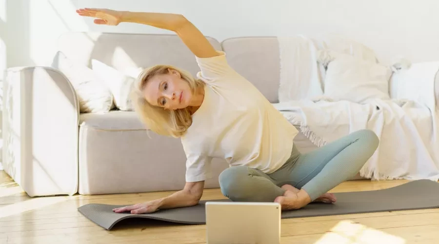 Middle-aged blonde woman, sitting on yoga mat in front of laptop doing stretches