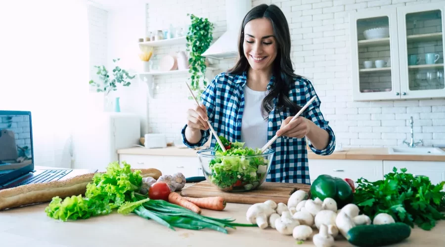 Woman in kitchen mixing a salad surrounded by various vegetables