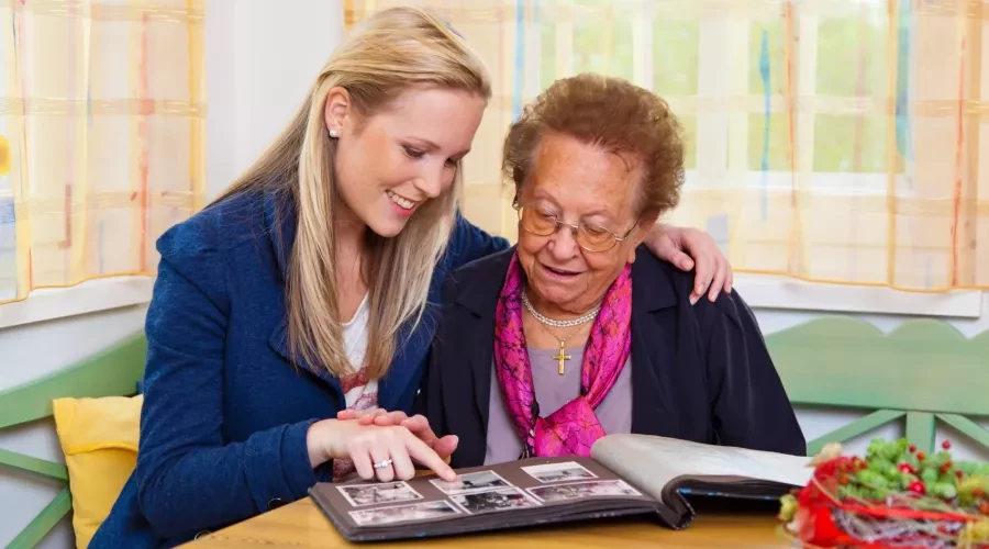 Young woman with arm around senior woman, both smiling and looking at photo album
