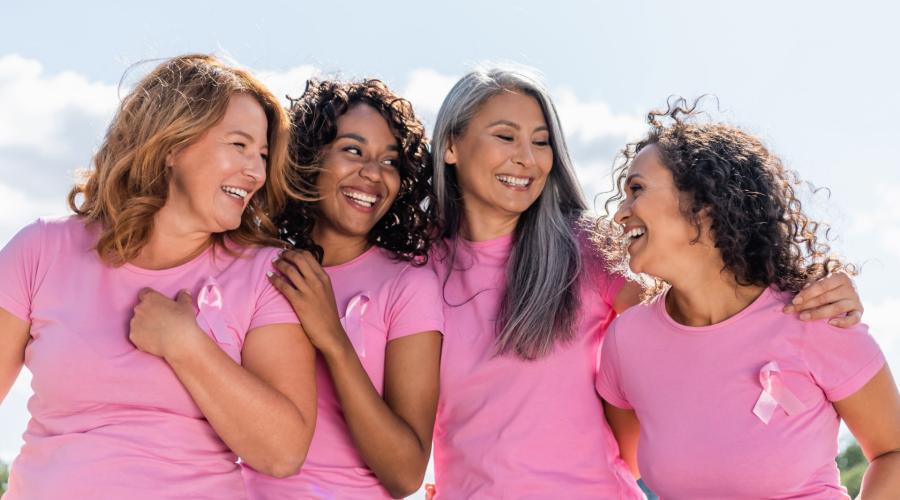 4 woman embracing and smiling in pink Breast Cancer awareness shirts with pink ribbon