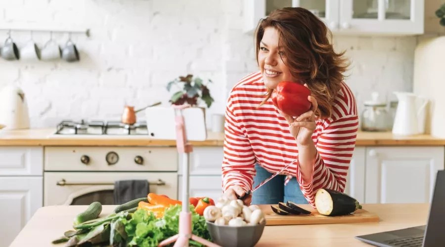 Middle-aged woman in kitched holding red bell pepper and cutting an eggplant on cutting board