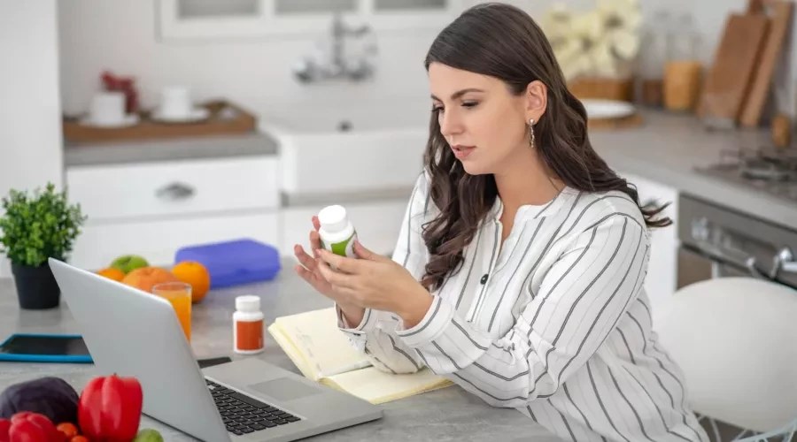 woman in kitchen with laptop looking at vitamin bottle
