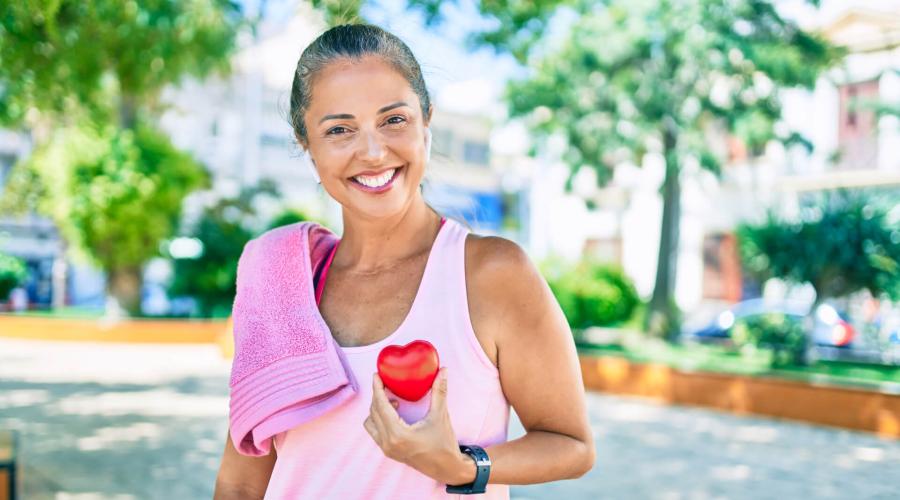Middle-aged woman outside with towel on shoulder and workout clothes, smiling and holding a heart
