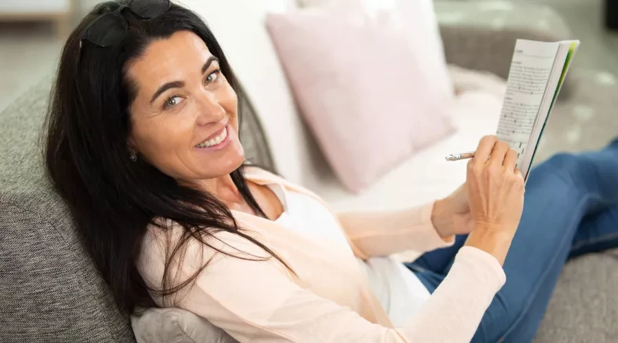 Woman on couch smiling and playing cross word puzzle in magazine