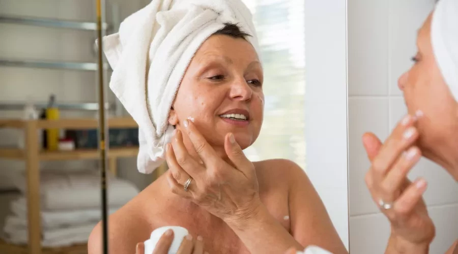 woman with towel on her head and looking in mirror, applying skincare product to her face after shower