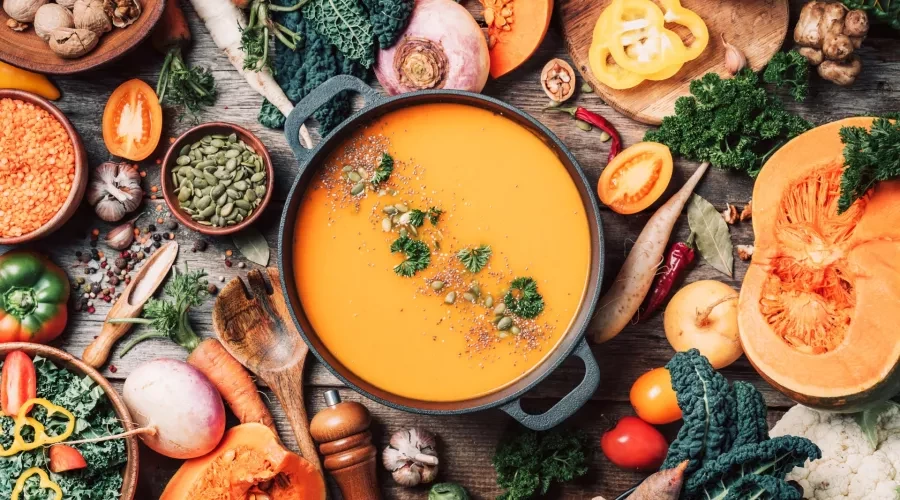 Iron pot with pumpkin soup on wood table, surrounded by vegetables and assorted nuts