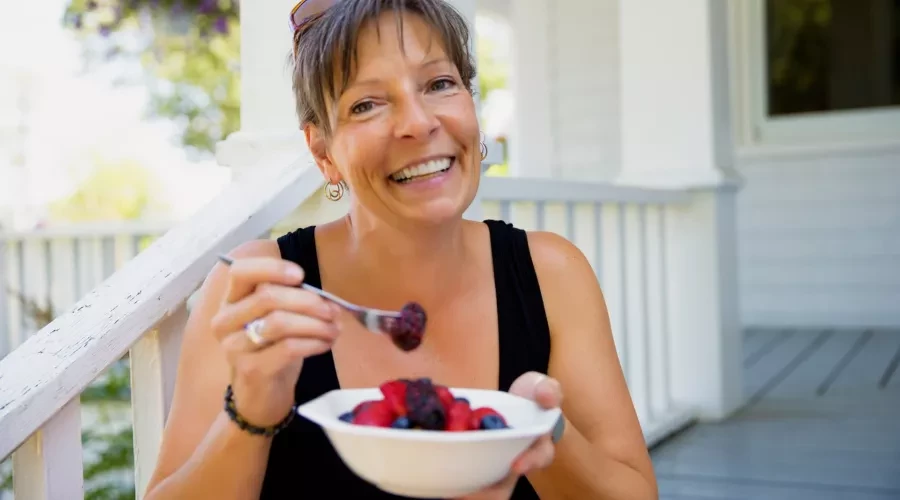 Woman smiling, eating a bowl of colorful fruit on front porch steps