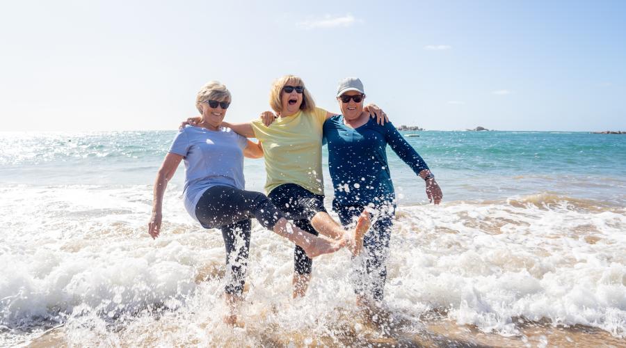 3 midlife women on ocean beach with arms around each other and kicking feet in water