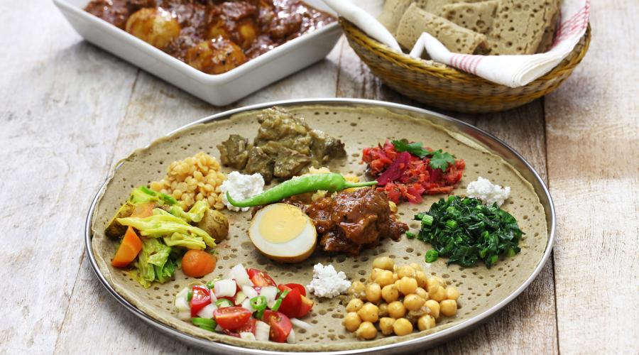 A plate of fresh, colorful Ethiopian food with adjacent dishes of meat stew and a basket of traditional flatbread.
