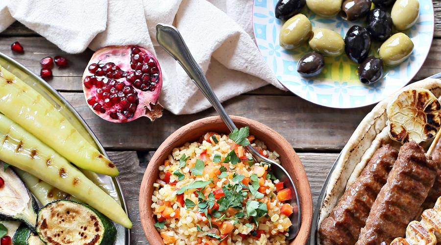A table of Mediterranean food including grain salad, olives, pomegranate, and grilled vegetables.