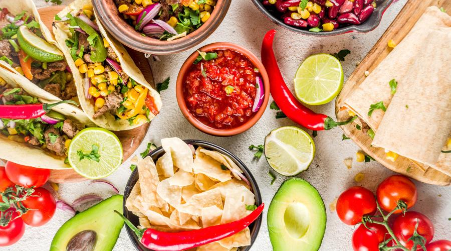 Colorful Mexican food including tacos, tortilla chips, salsa, avocados, limes and other produce laid out on a table.