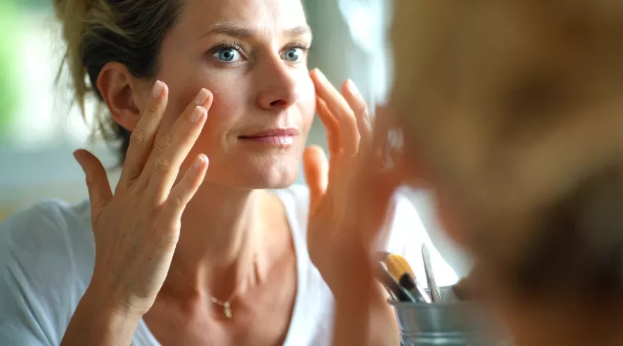 A woman looks in the mirror and carefully applies a skincare product to her cheekbones.