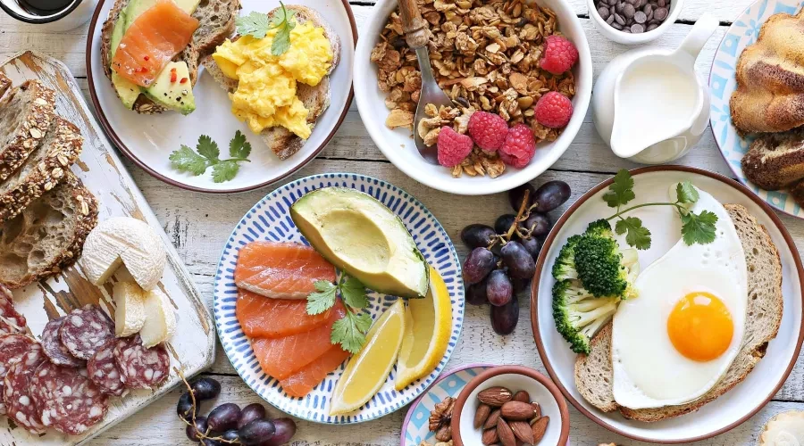Wood table with bowls of food including Granola and berries, eggs on toast, cheese and salami, nuts and cake