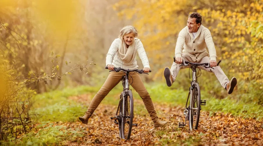 An older man and woman riding bikes through fall foliage with their feet raised playfully off the pedals.