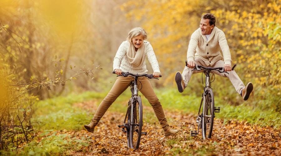 An older man and woman riding bikes through fall foliage with their feet raised playfully off the pedals.