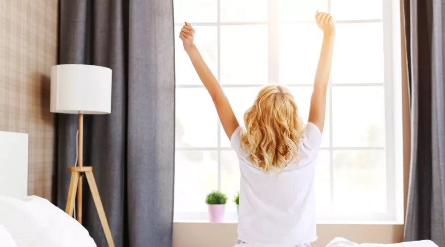 Women stretching with arms in the air, sitting on bed staring out window with sunlight