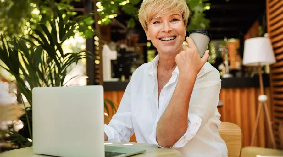 Professional woman sits outdoors working at her laptop with a coffee and smiles into the camera.