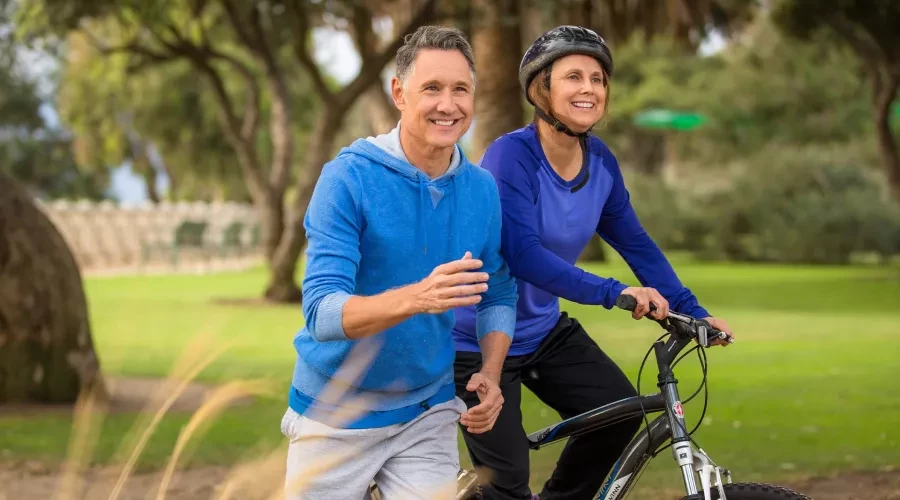 Couple in 50's in park, man with blue sweatshirt running & woman in blue shirt on her bike
