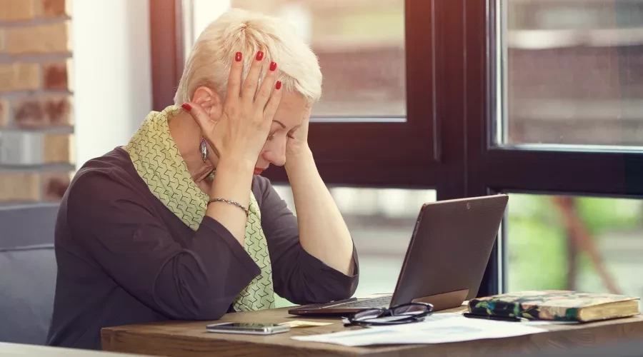 Middle aged woman at desk with laptop, stressed with hands on her head