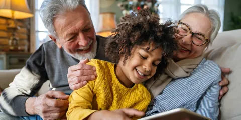 Senior couple on couch with kid in lap looking and laughing with ipad, christmas tree in background