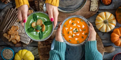 Womens hands holding bowls of vegetable soup over a wood table displayed with pumpkins, vcrackers, spices and nuts