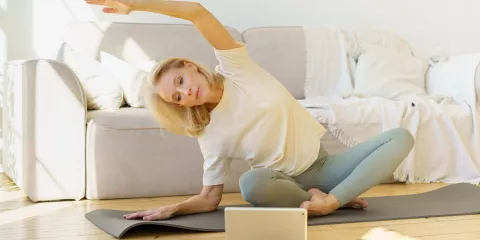 Middle-aged blonde woman, sitting on yoga mat in front of laptop doing stretches
