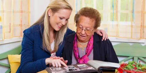 Young woman with arm around senior woman, both smiling and looking at photo album