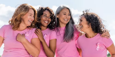 4 woman embracing and smiling in pink Breast Cancer awareness shirts with pink ribbon