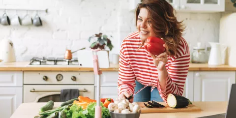 Middle-aged woman in kitched holding red bell pepper and cutting an eggplant on cutting board