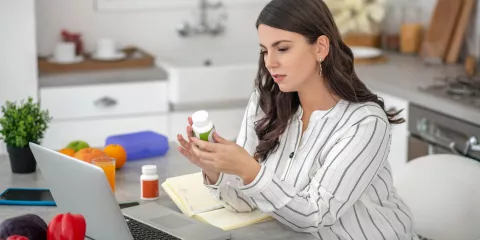 woman in kitchen with laptop looking at vitamin bottle
