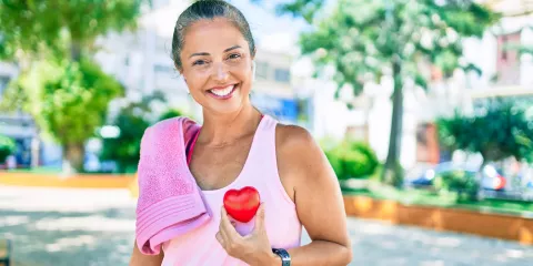 Middle-aged woman outside with towel on shoulder and workout clothes, smiling and holding a heart
