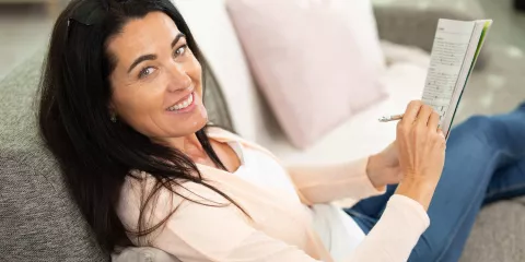 Woman on couch smiling and playing cross word puzzle in magazine