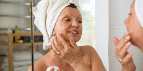 woman with towel on her head and looking in mirror, applying skincare product to her face after shower