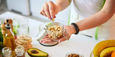 Woman in kitchen with jar of cashews in her hand, avocado, spices, walnuts, oil on the table 