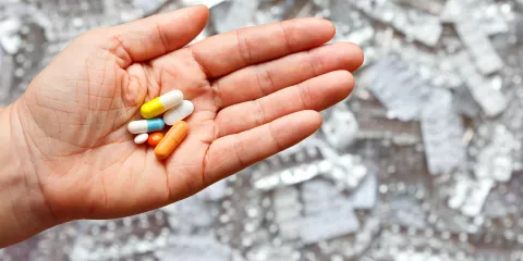 Pills and capsules in a person's open palm with a pile of empty pill blister packs out of focus in the background.